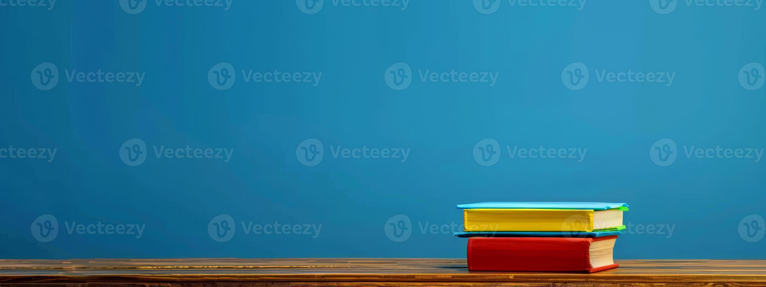 A stack of vibrant books in various colors is placed on a wooden table, creating an inviting atmosphere in a warm and cozy space with a blue backdrop photo