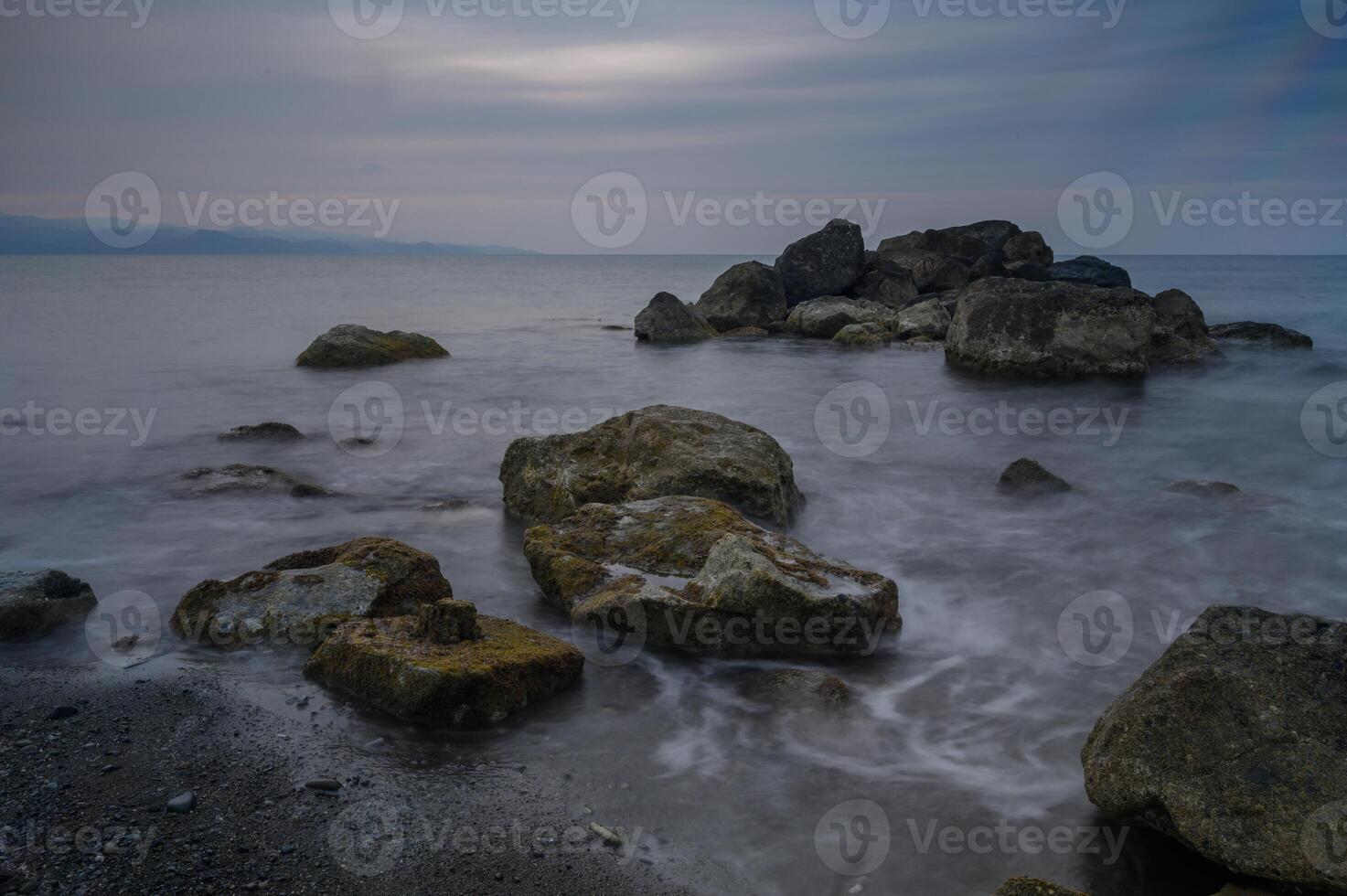 Calm waves gently lap against rugged stones on a serene shoreline at dusk near photo