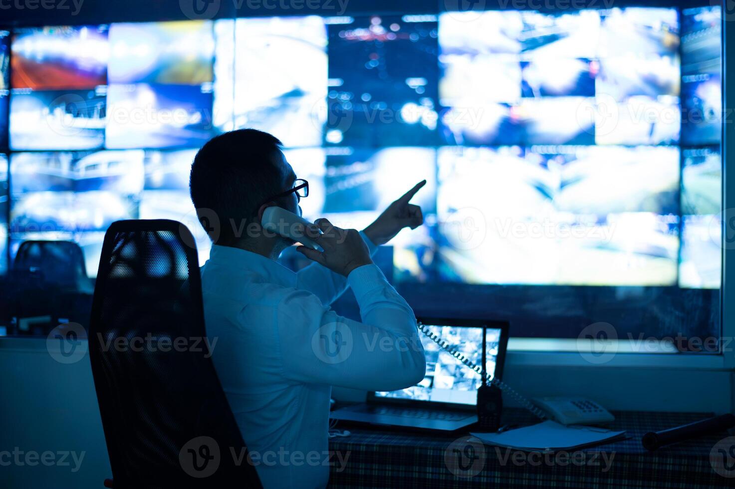 The CCTV operator is talking on the phone and pointing at the monitor to check the image on the screen. photo