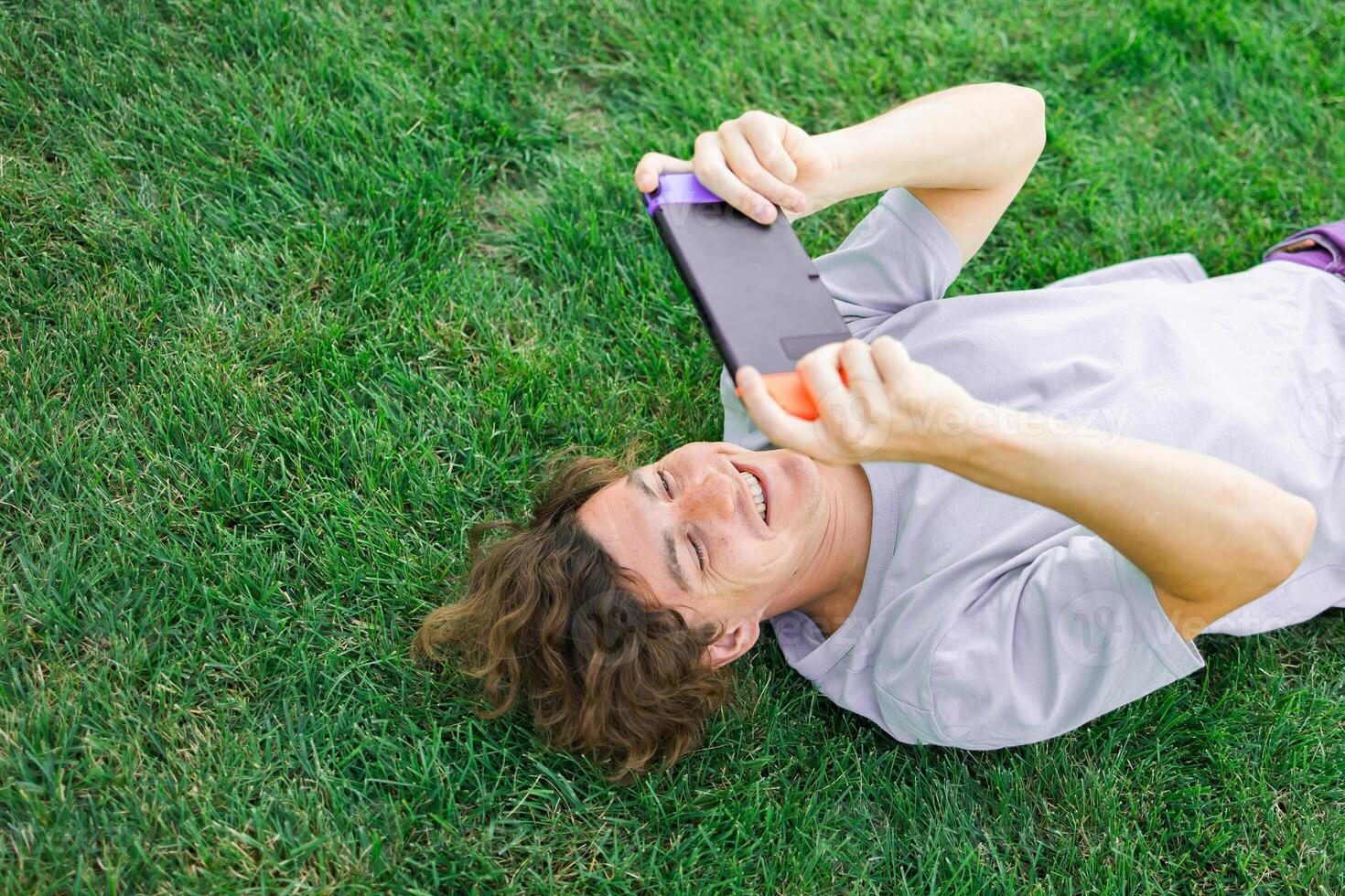 Young man smiling confident playing game at park lying on a grass. Technology and gaming concept photo