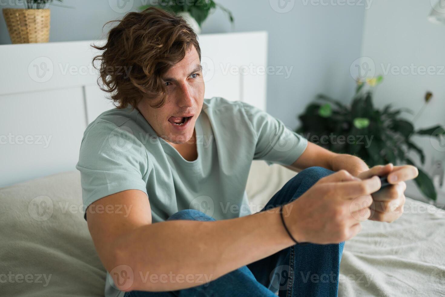Handsome excited gamer sitting on a bed and playing games on a console. Wireless controller and online game technologies concept photo