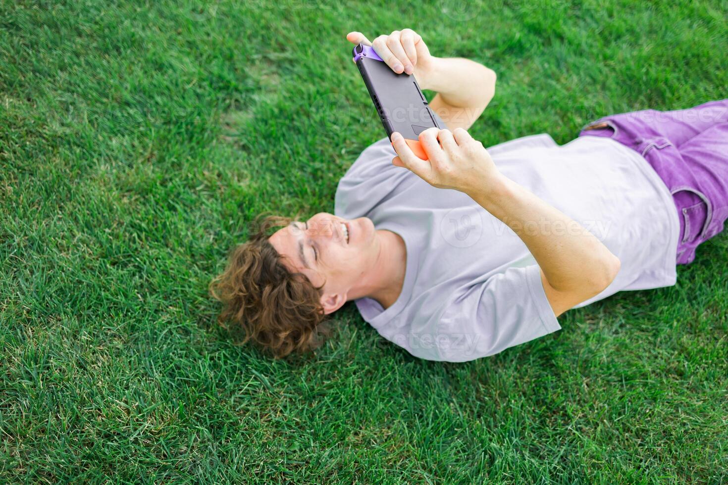 Young man smiling confident playing game at park lying on a grass. Technology and gaming concept photo