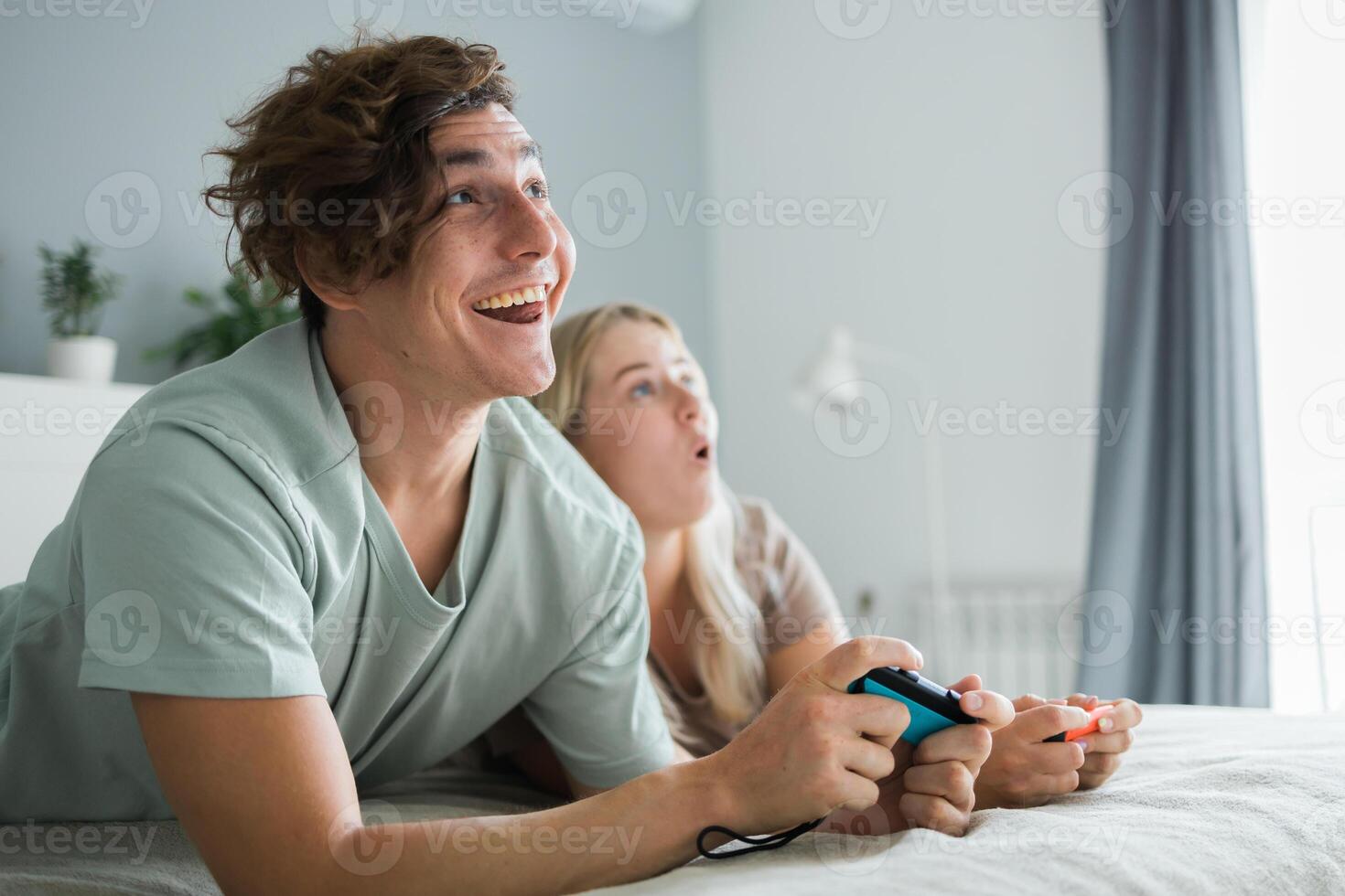 Young couple in love playing games while lying on the bed in the bedroom, having fun and enjoying each other on their weekends. Technologies and gaming concept photo