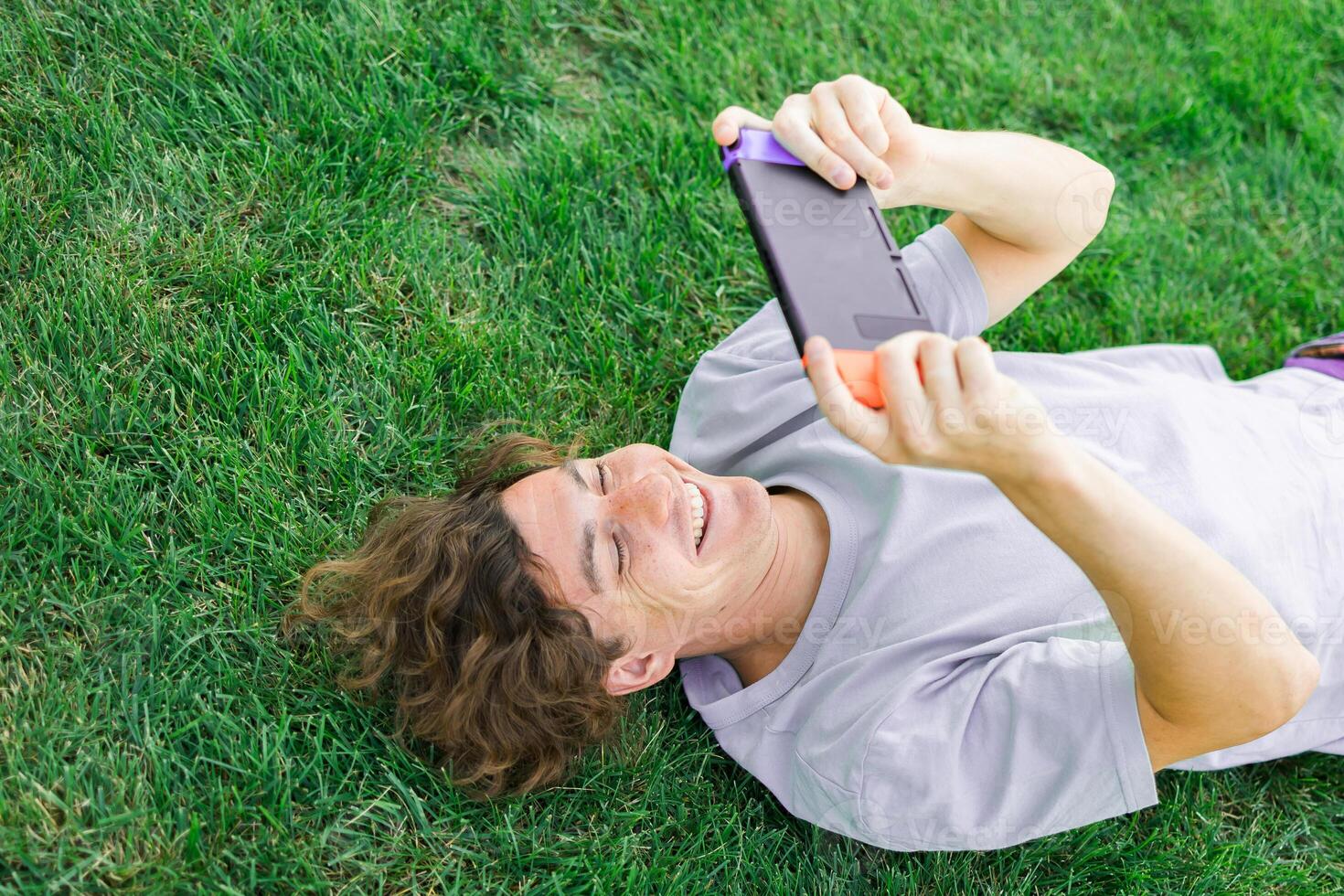 Joyful man smiling and having fun while playing games with portable console and lying on the grass outdoors. Technology and game addiction concept photo
