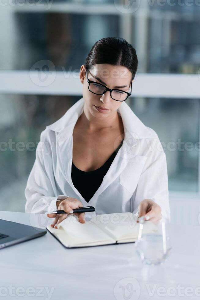Caucasian mixed-race businesswoman in glasses writing notes while sitting at desk in the office. People and wireless technology concepts. Working on laptop writing letter in notebook. Vertical photo