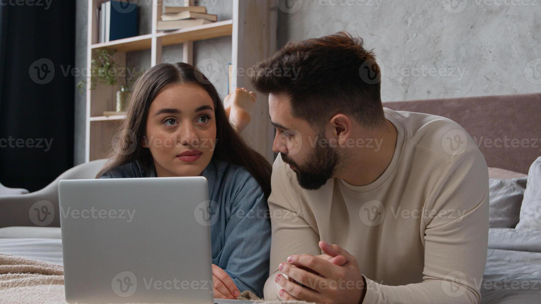 Caucasian married couple man woman talking discuss shopping online on laptop at home young family boyfriend girlfriend buying goods internet store looking at computer lying on bed ordering purchase photo