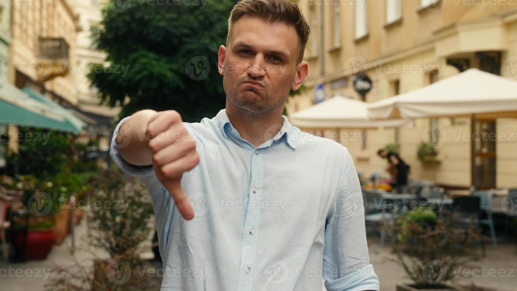 Sad frowning man upset displeased in city looking at camera thumb down finger refusal dislike gesture male businessman tourist outdoors sign negative rejection disapproval business guy reaction bad photo