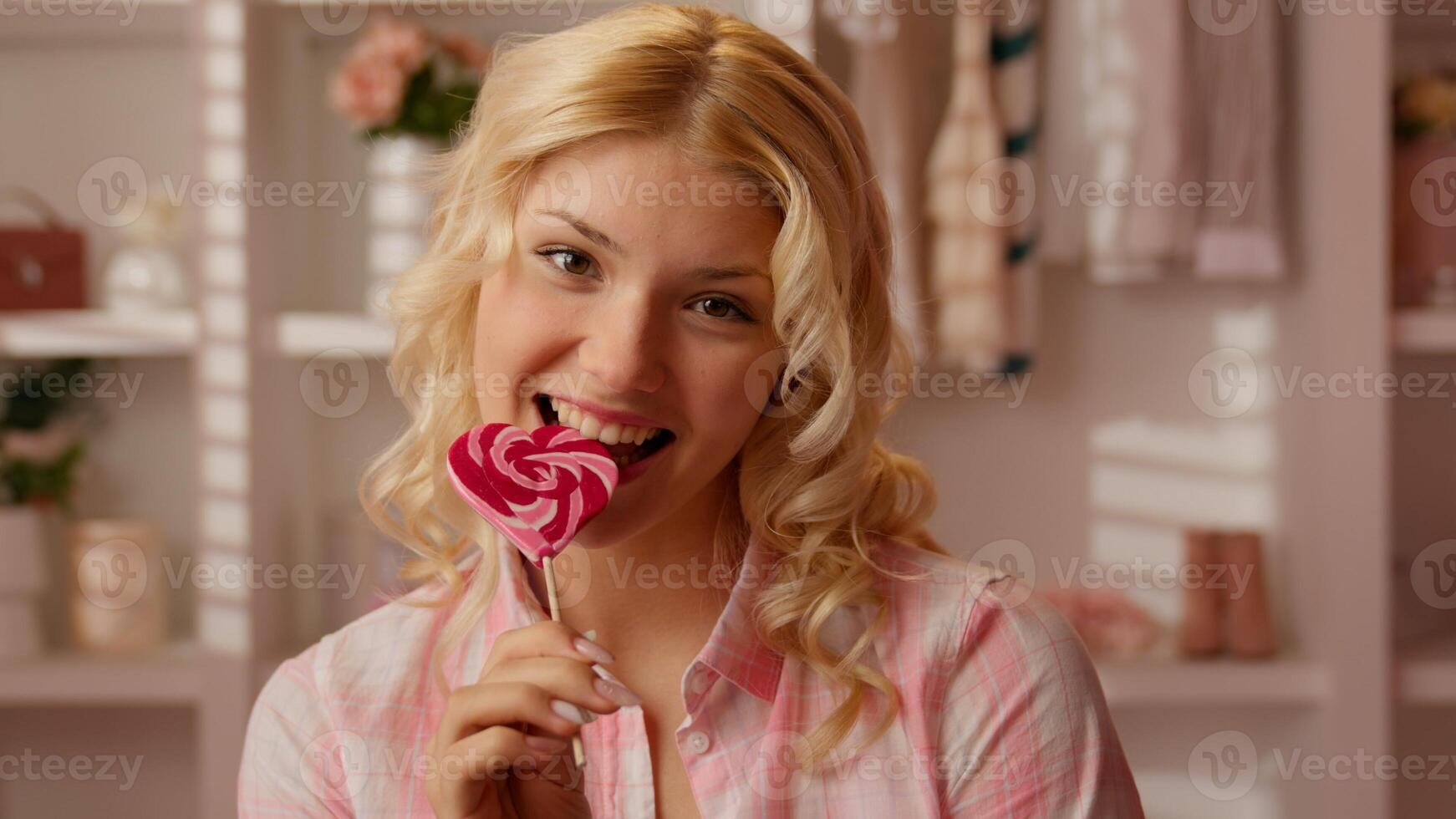 Joyful happy Caucasian woman teen girl teenager female in glamour bathroom smiling bite pink sweet caramel lollipop in shape of heart on wooden stick candy delicious dessert food nutrition overweight photo
