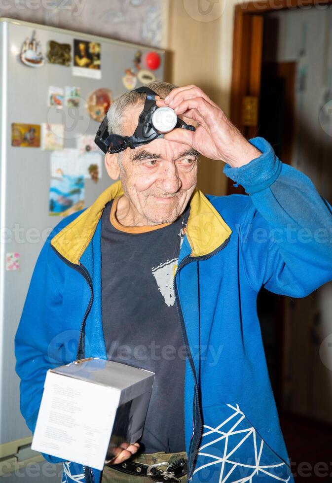 A 76 year old Russian man smiles as he tries on a new headlamp he received as a gift. Holding the box in his hands, he stands in a cozy rural home interior, radiating joy and warmth photo
