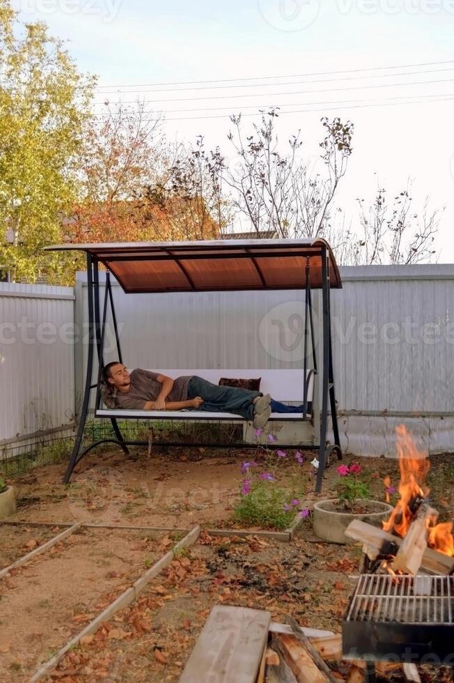 Sleeping young man laying on a garden swing next to bonfire photo