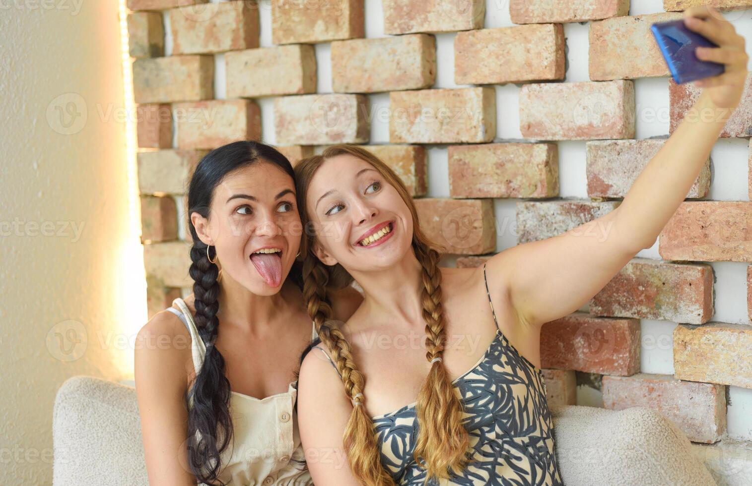 Ukrainian woman brunette and Russian woman blonde portrait together with braids posing and taking a selfie, showing tongue photo