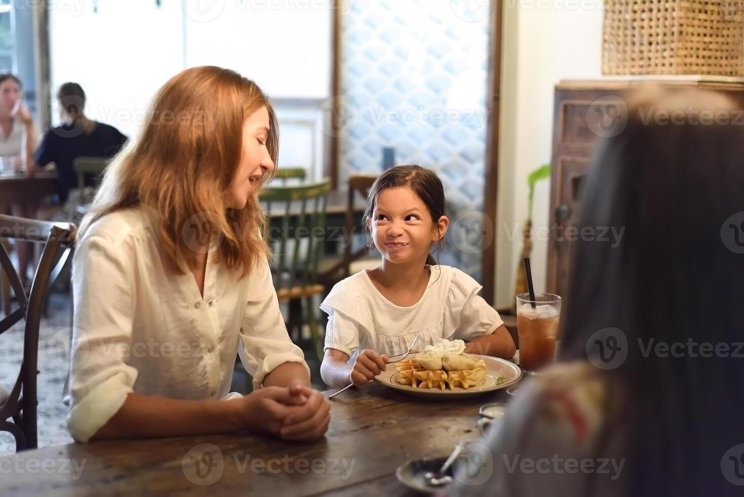 Little girl and her mother eat sweet belgian waffles with banana and cream in a cafe, daughter making a funny face photo