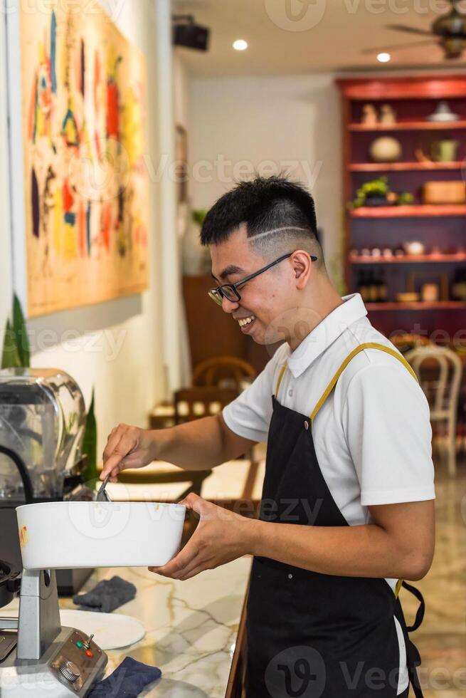 Vietnamese man chef in uniform holds spoon and bowl with liquid dough for belgian waffles photo