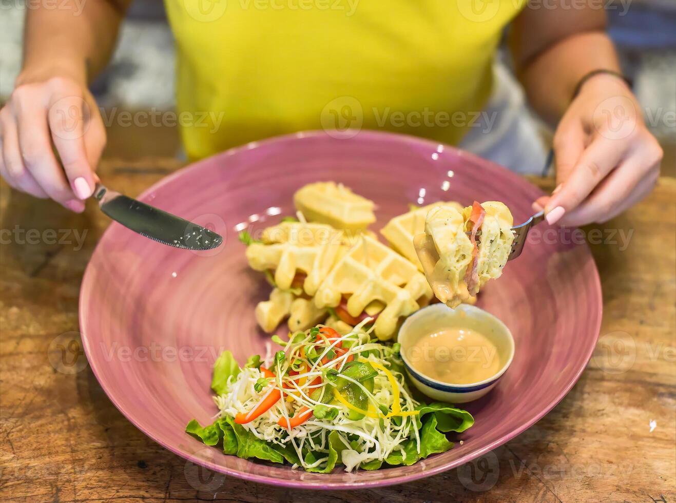Person eating belgian waffles with ham and cheese and salad, holding fork and knife in hands photo