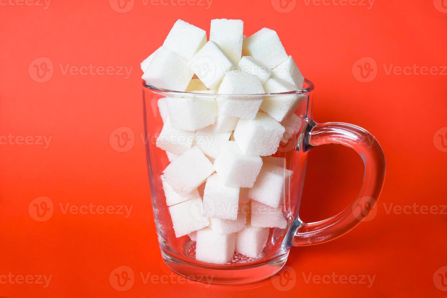 Transparent mug full of refined white sugar cubes on red background photo
