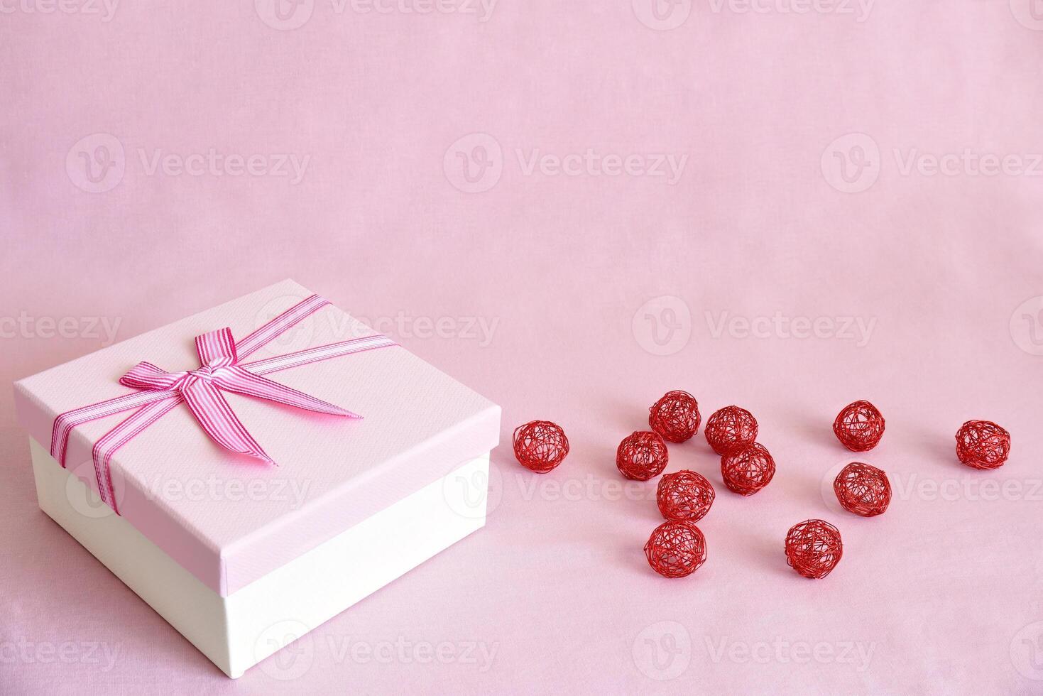 White gift box with pink lid and bow next to red metal balls on pink fabric background with copy space. Celebration concept photo