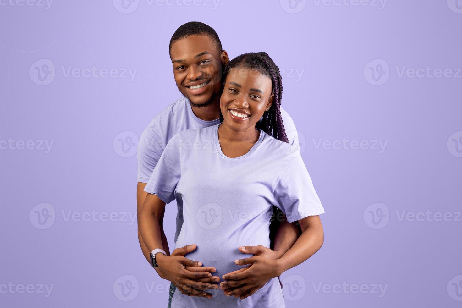 Portrait of smiling black guy hugging pregnant wife from back, cheerful couple standing over purple violet studio background, thinking about baby's name or gender. Empty speech bubble, free copy space photo