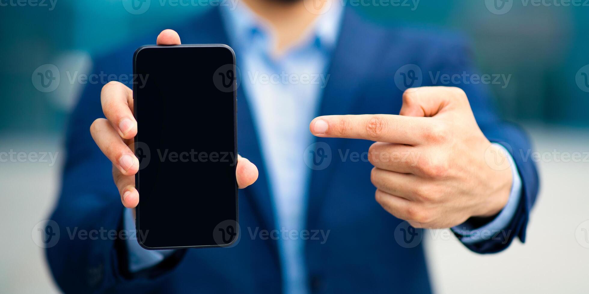 Modern technologies and business concept. Unrecognizable businessman in stylish suit pointing at smartphone with blank screen, recommending brand new mobile application for business, finance, closeup photo