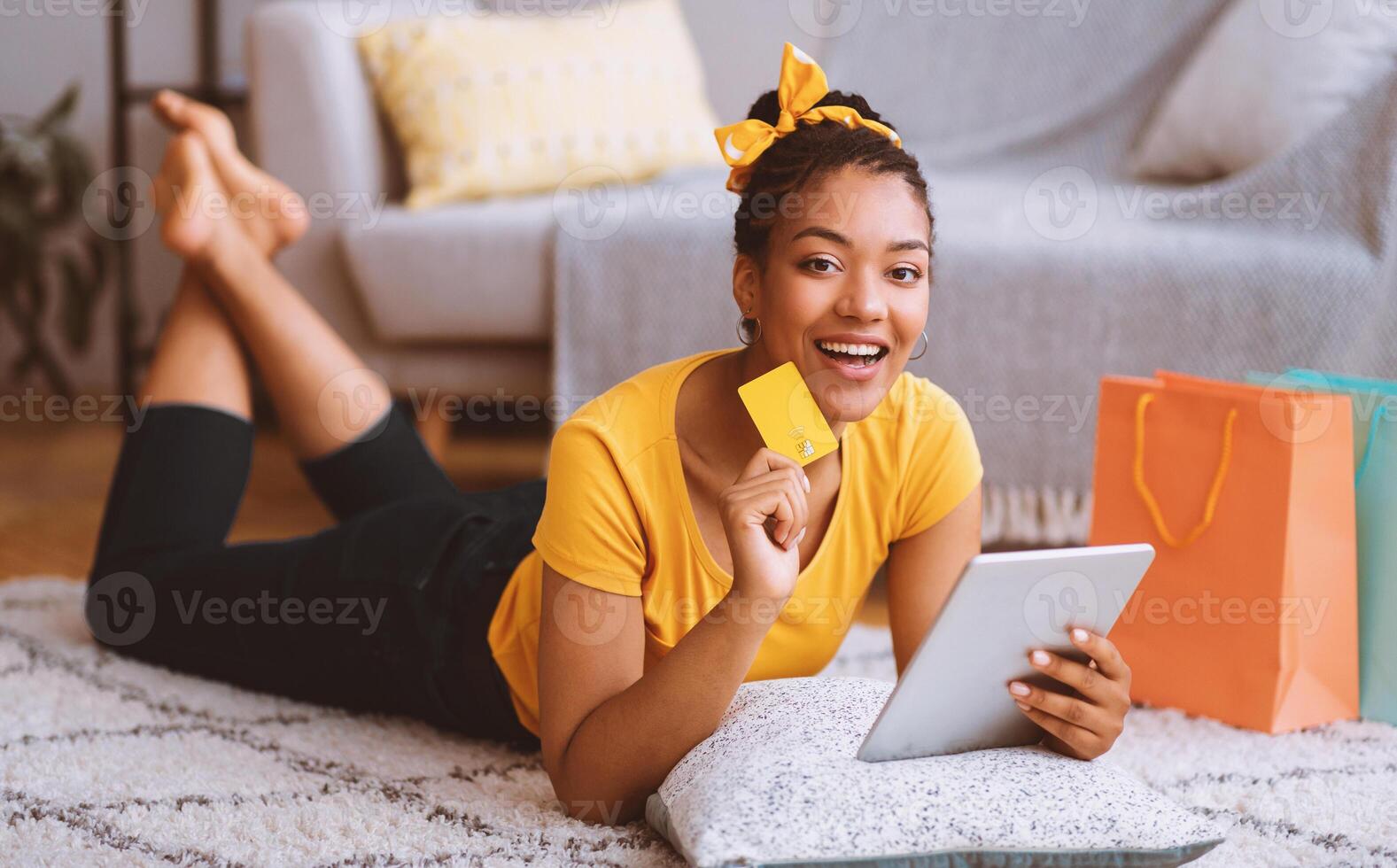 Online Shopping Concept. Happy Black Woman Using Digital Tablet, Holding And Showing Credit Card, Buying And Ordering Things In Internet Shop Lying On The Floor Carpet, Looking At Camera photo