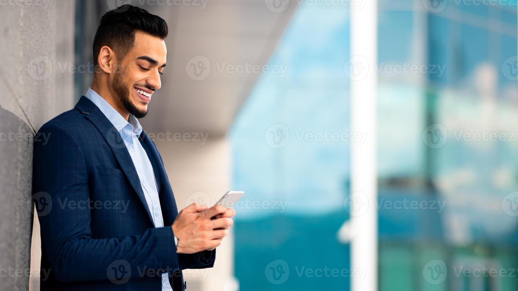 Handsome middle eastern businessman in suit standing by modern business center, using cellphone and smiling, sending emails while having break, panorama with copy space, side view photo