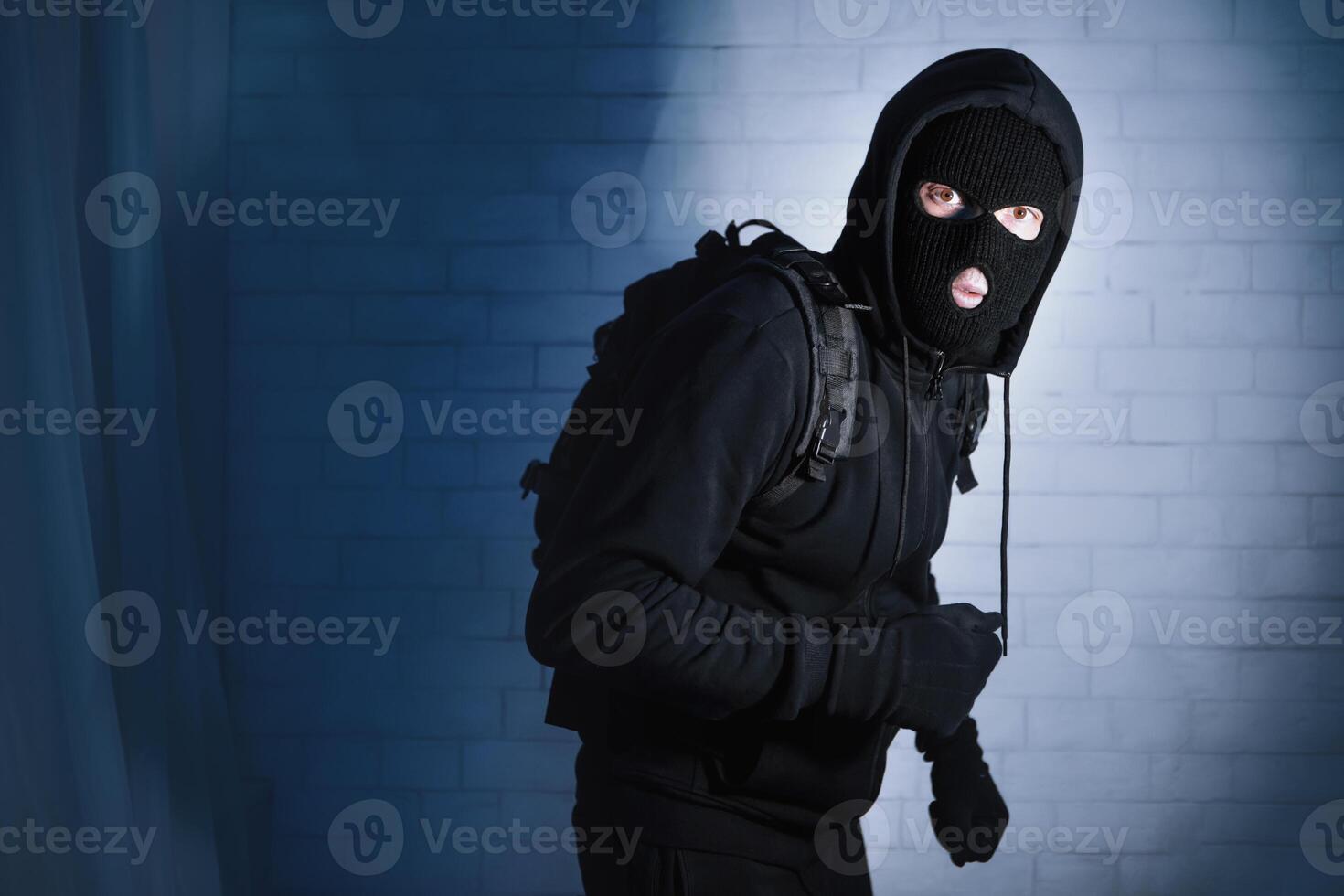 Burglary Concept. Intruder in black balaclava standing in apartment or office in the dark, looking at camera photo