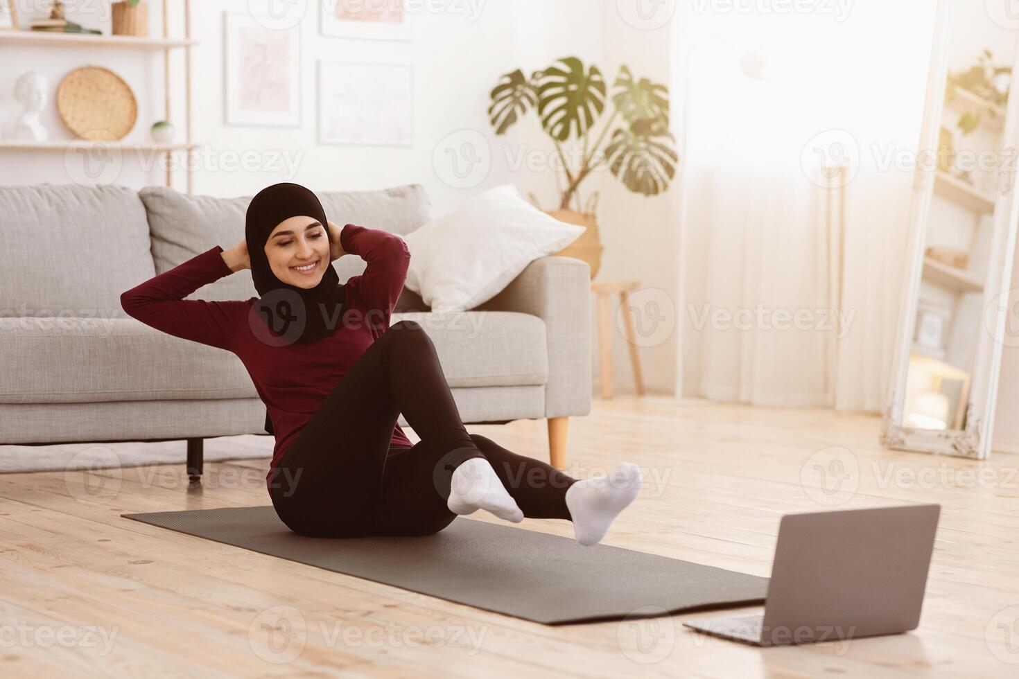 Sporty arabic woman in hijab doing abs exercises with laptop at home, watching online tutorials, enjoying healthy lifestyle, copy space photo
