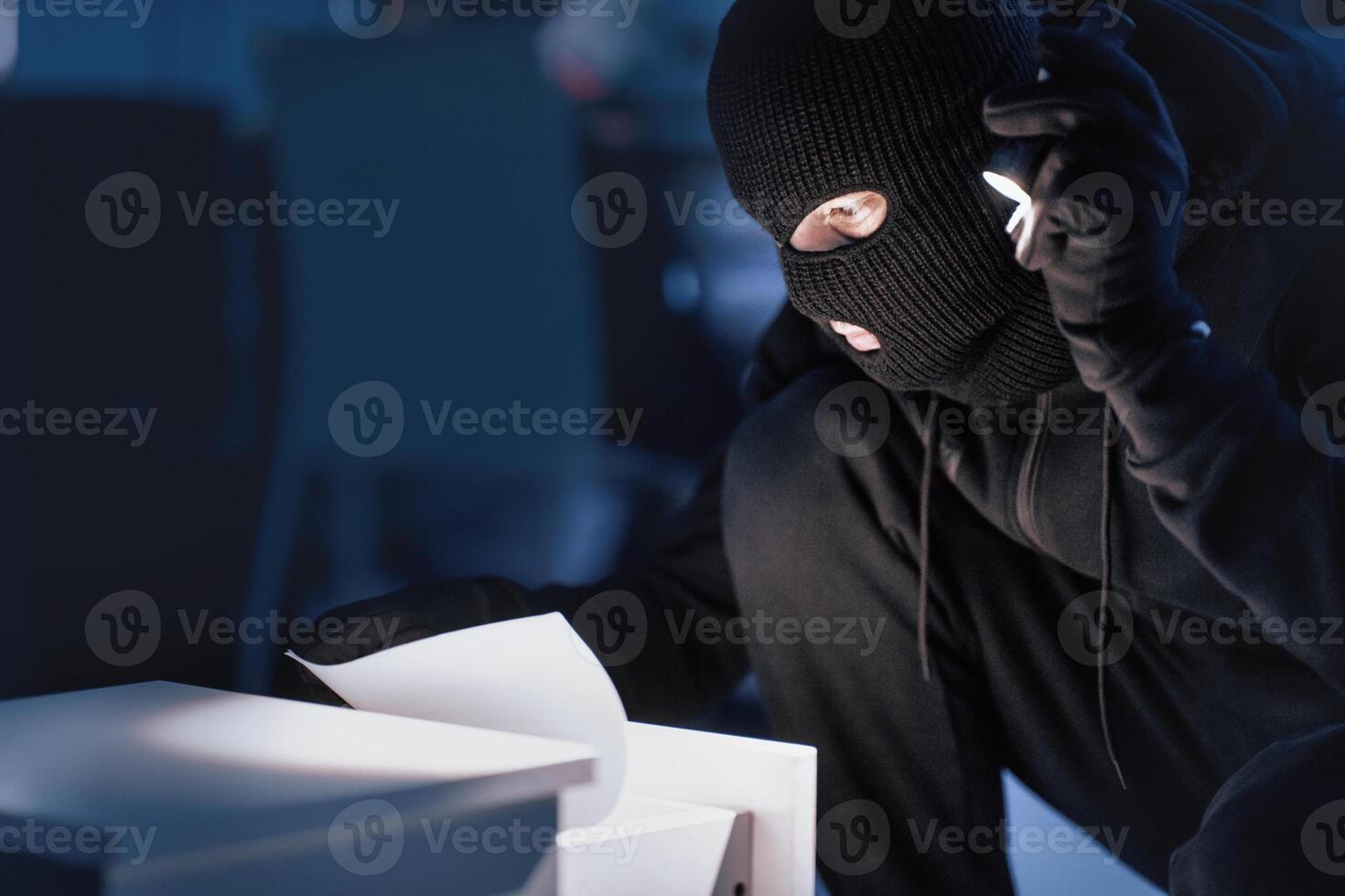 Closeup of disguised robber with flashlight searching for documents in office at night, copy space, blurred background photo
