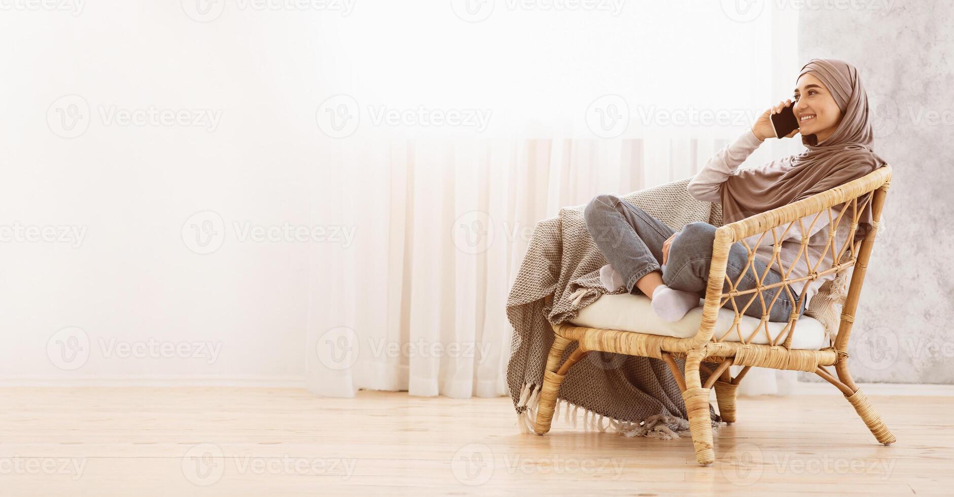 Muslim girl in hijab talking on cellphone while relaxing in cozy chair at home, sitting against window, side view with copy space photo
