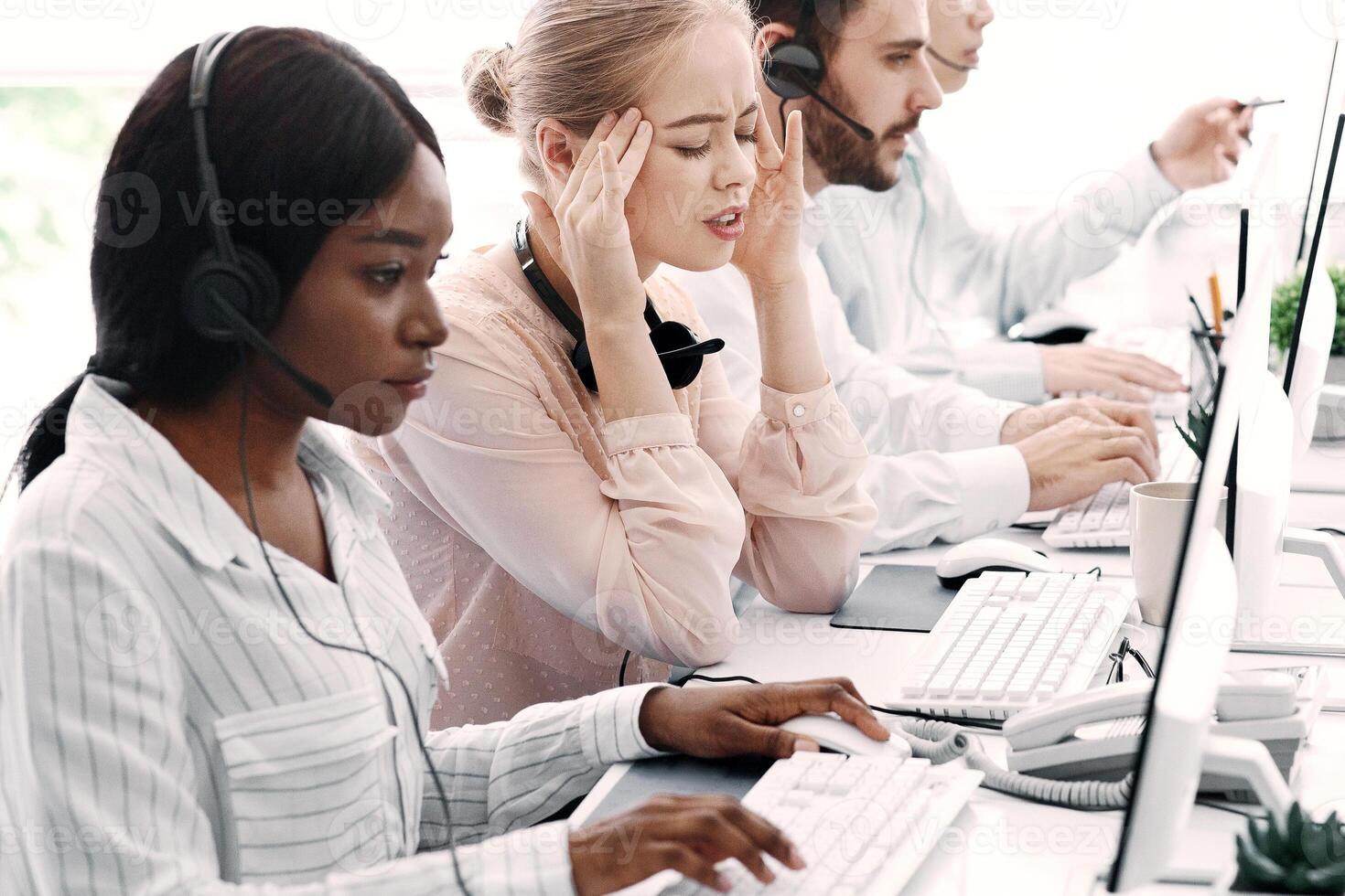 Technical support operator suffering from headache and her colleagues working with computers at office photo
