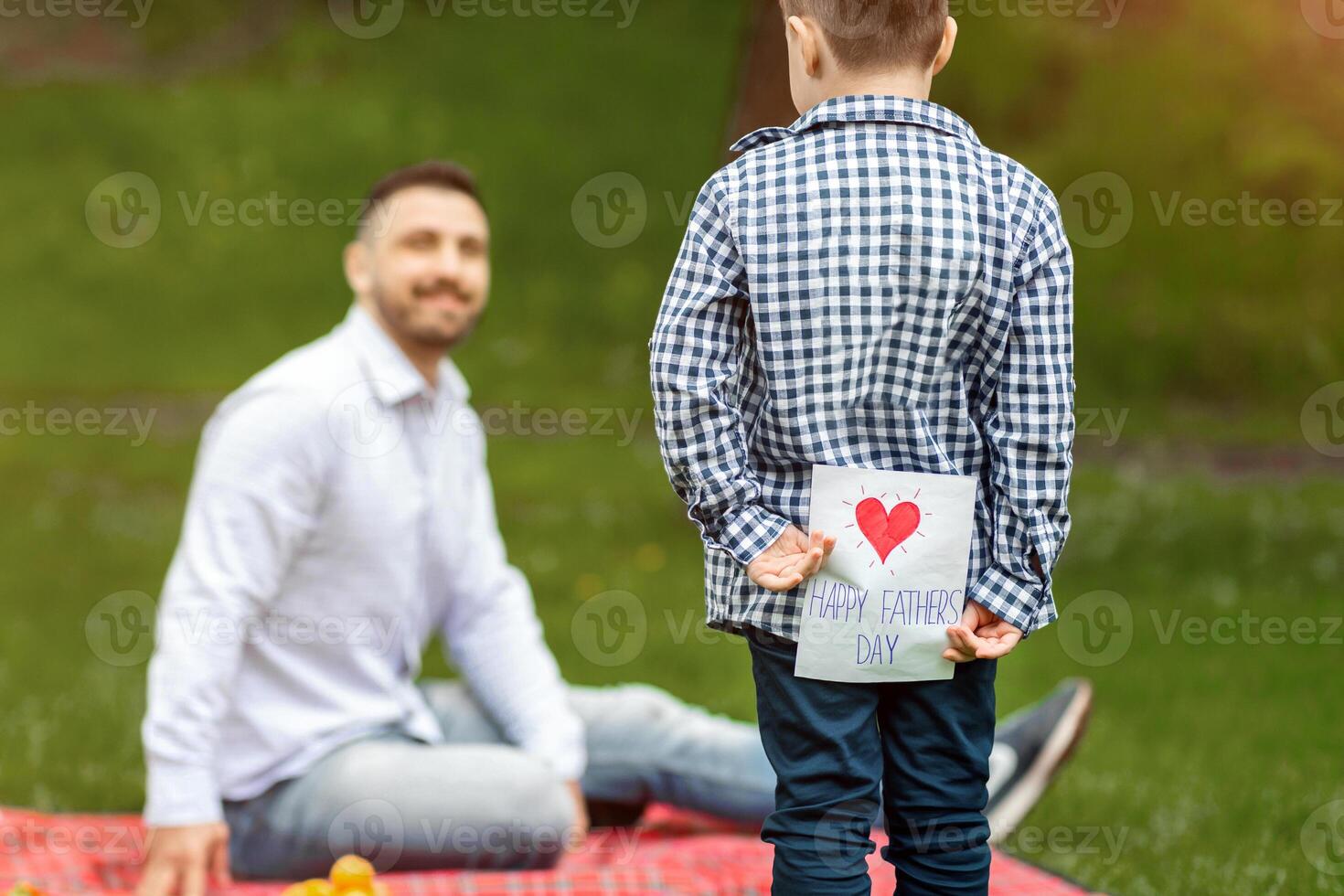 Father's Day surprise. Child hiding greeting card for dad behind his back on picnic in park, empty space photo