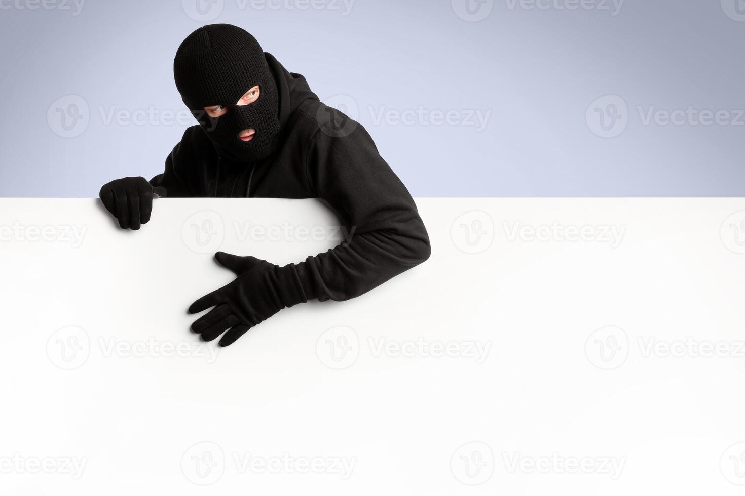 Masked gangster holding white blank advertising panel and looking back, isolated over white studio wall photo