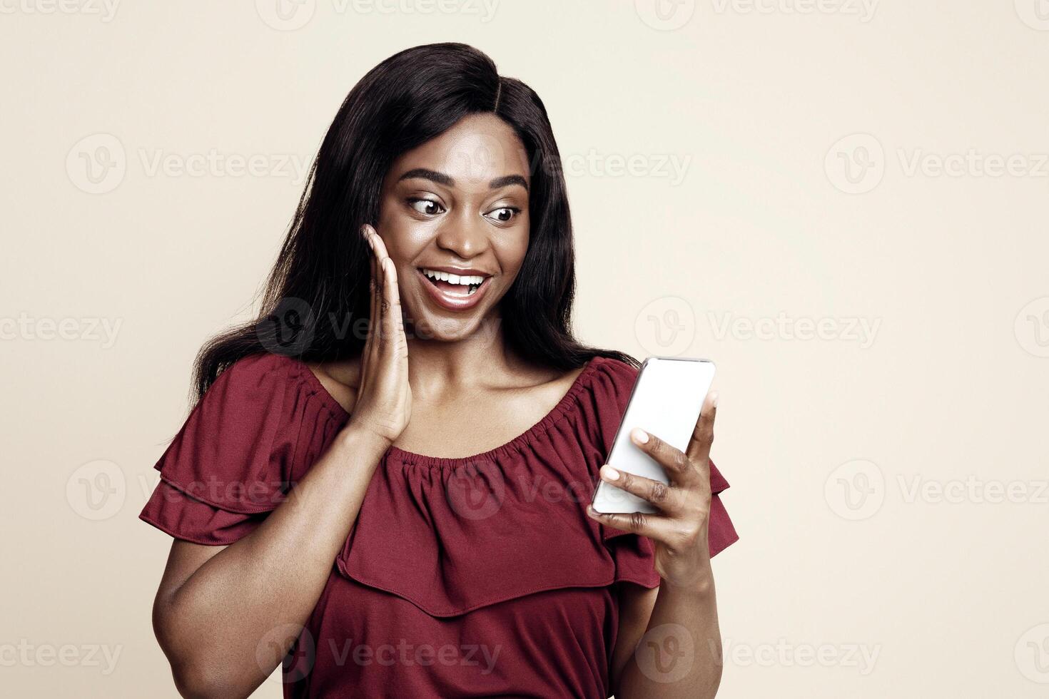 Emotional stylish millennial black woman using nice mobile application on brand new smartphone, touching her cheek, surprised with newest shopping app, beige studio background, copy space photo