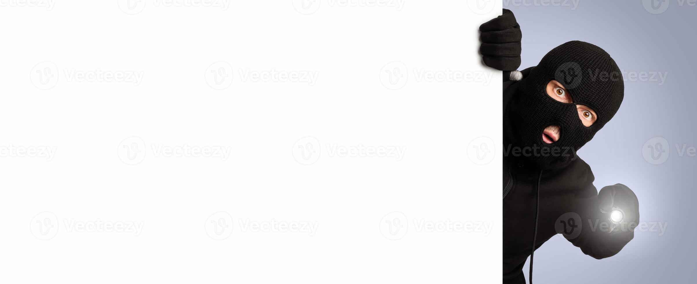 Surprised criminal wearing black balaclavausing torch and peeking out white panel isolated on gray studio background photo