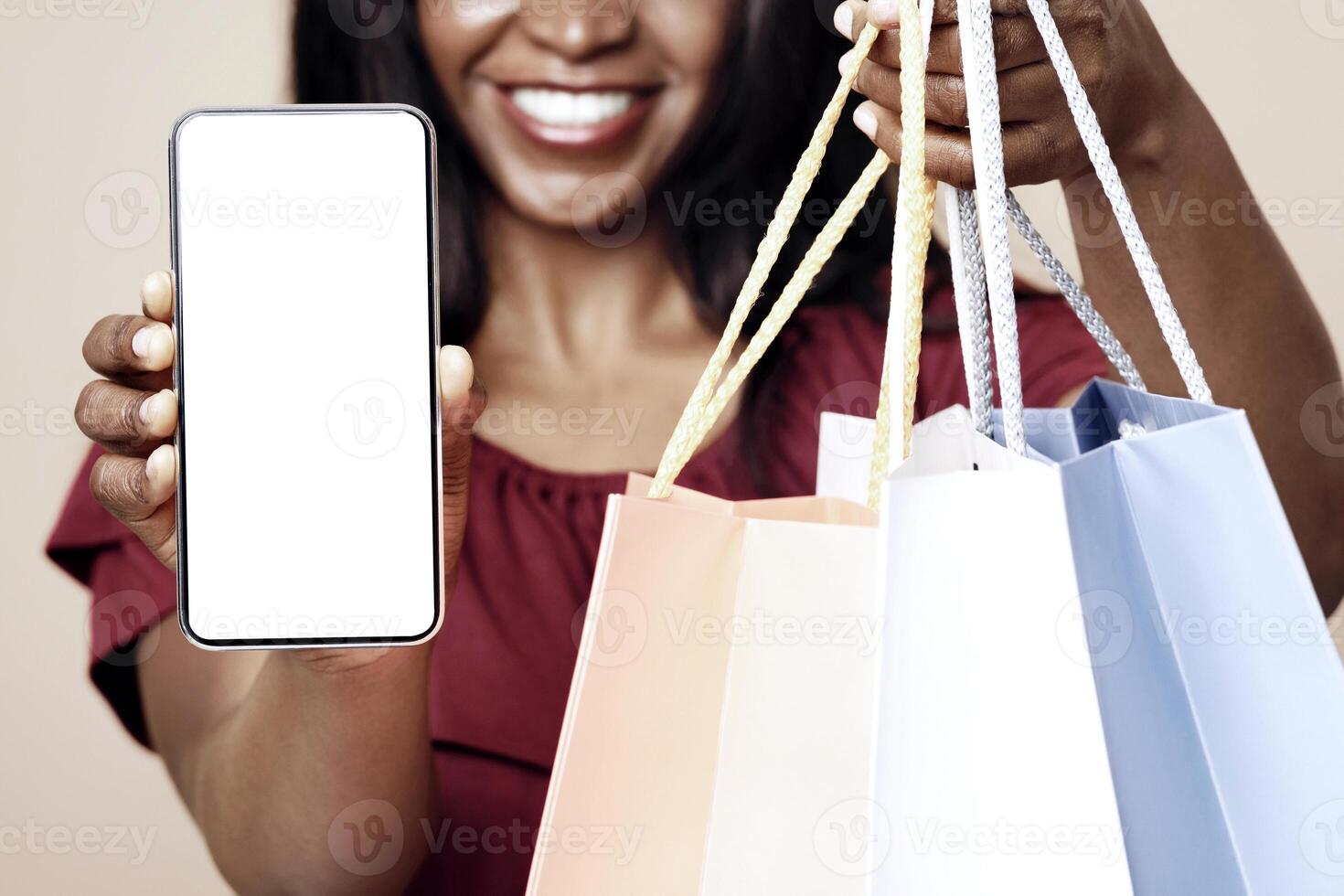 Cropped of african american woman shopaholic showing modern smartphone with empty screen and holding bunch of colorful shopping bags, unrecognizable lady recommending shopping mobile app, mockup photo