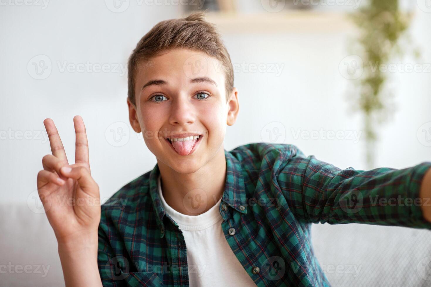 Handsome teen boy taking selfie, showing tongue and peace gesture, sharing in social media, home interior, teen lifestyle photo