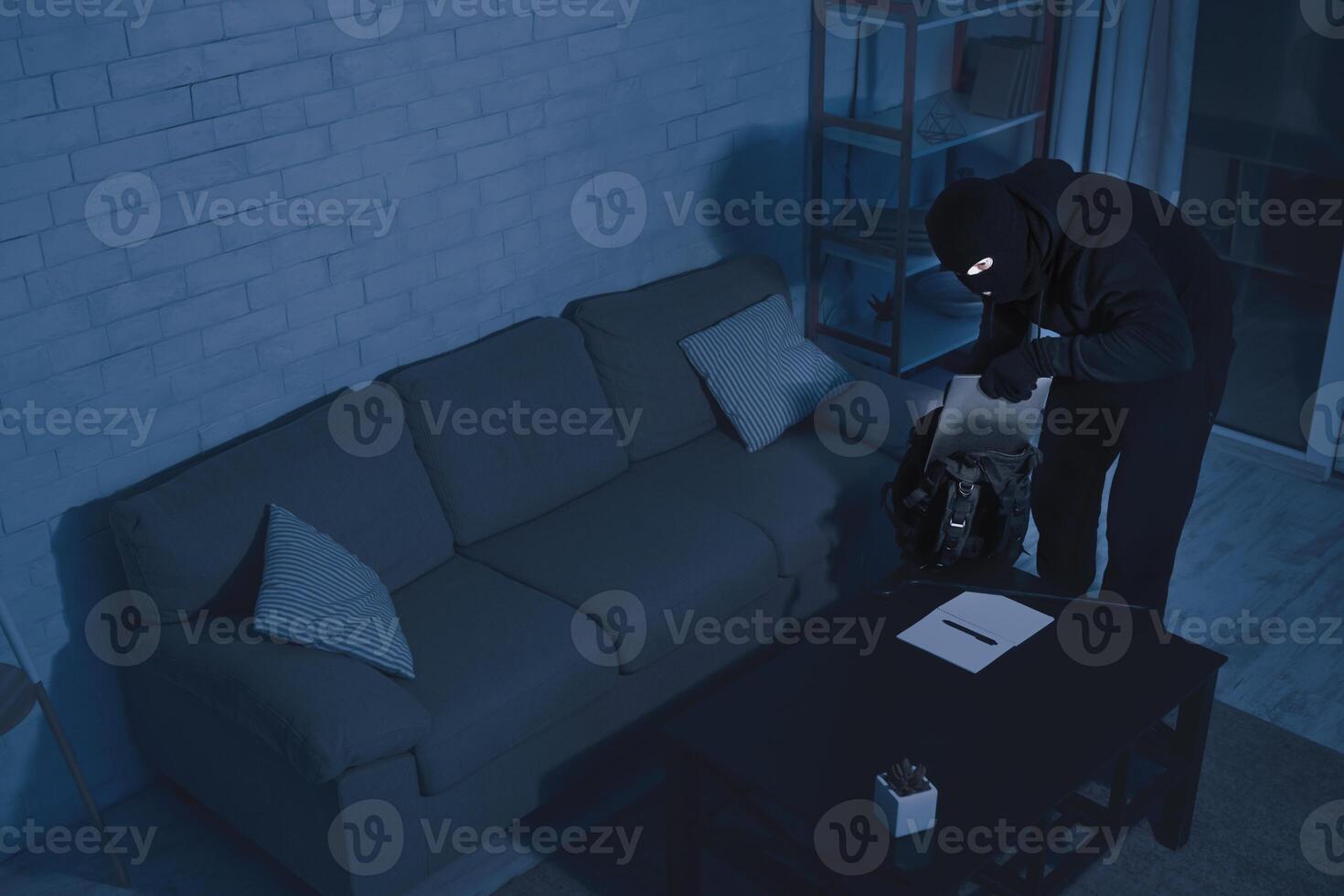 Security Camera Concept. Disguised thief stealing pc, putting it in his backpack in the night, high angle top view photo