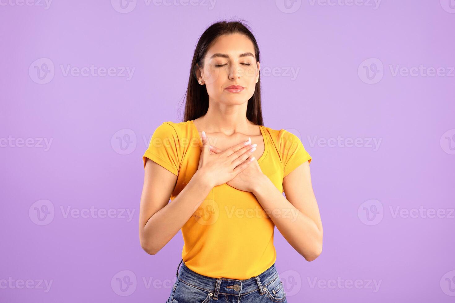 Thank You. Grateful Young Woman With Closed Eyes Keeping And Pressing Hands To Her Chest Expressing Gratitude Posing Over Purple Studio Background. Appreciation Concept photo