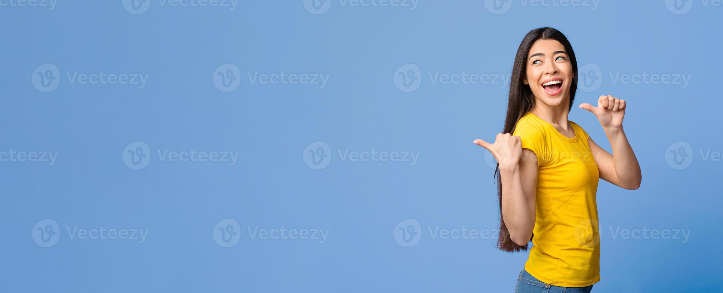 Best Choice. Joyful asian girl pointing on copy space behind her back with thumb ups, purple background, panorama photo