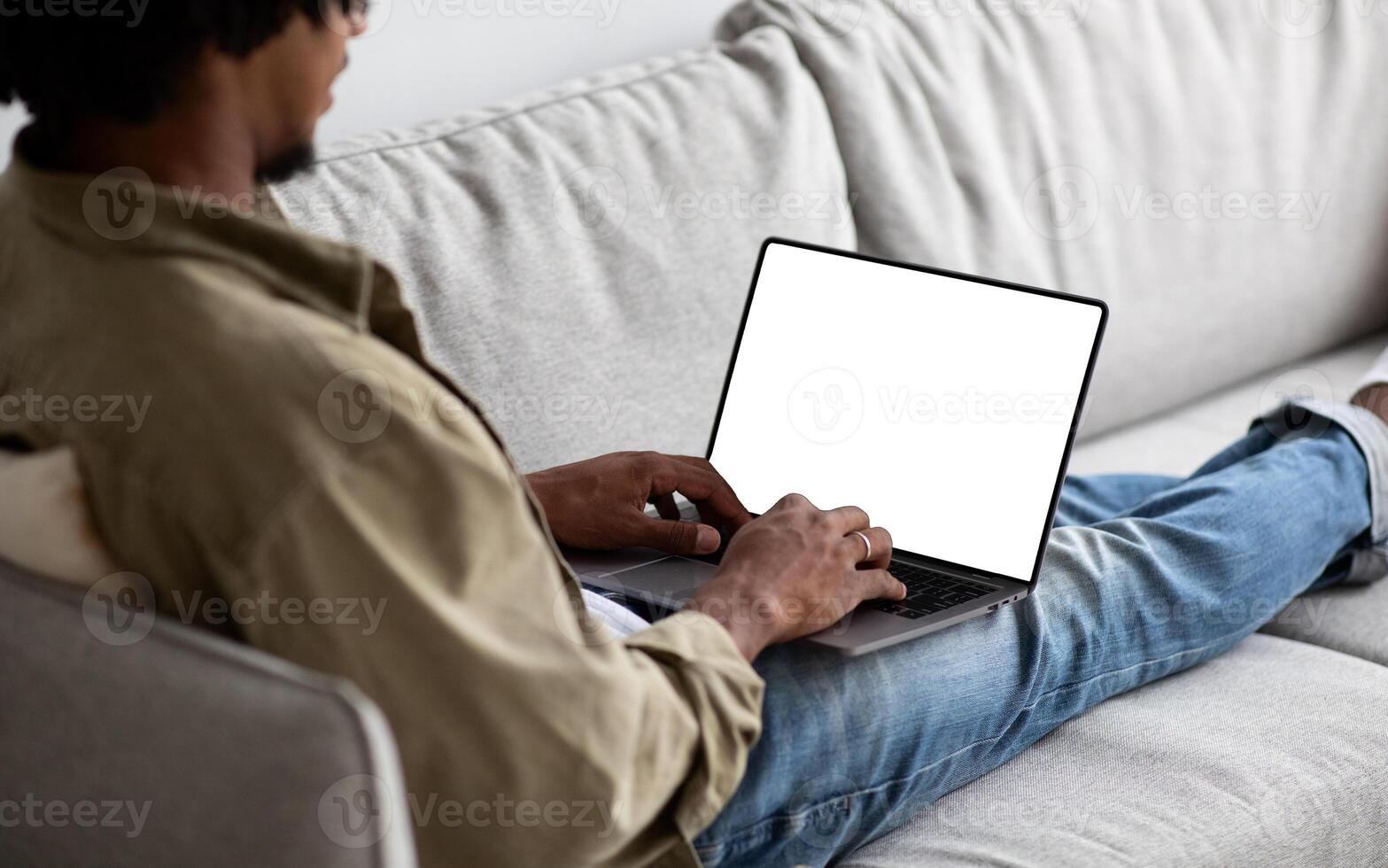Black Man Using Laptop Computer With Blank White Screen While Sitting On Couch At Home, African American Guy Working Online Or Browsing Internet Website, Over Shoulder View, Mockup With Copy Space photo