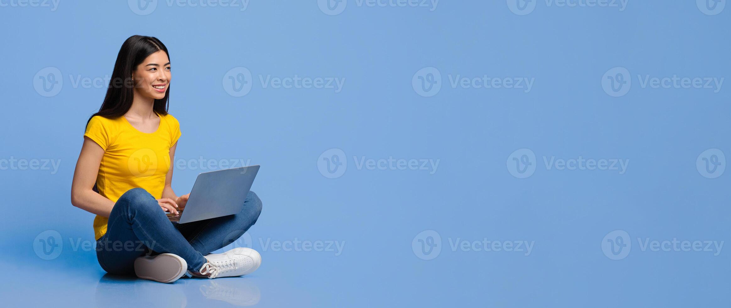 Smiling young asian woman sitting on floor with laptop computer over purple studio background, looking away at copy space, millennial korean lady studying or working with notebook photo