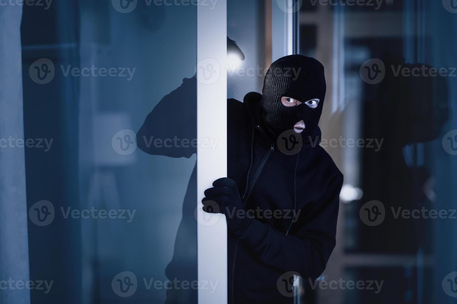 Is Anybody Home. Cautious masked thief looking and peeping into office through window or glass door with torch flashlight photo
