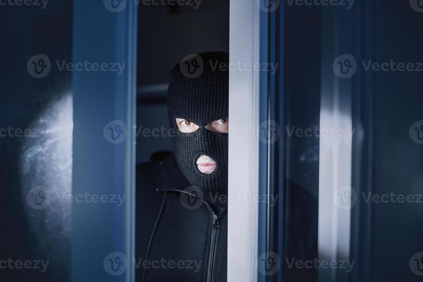 Burglary Concept. Evil thief looking at camera through open front door, entering enclosed property photo