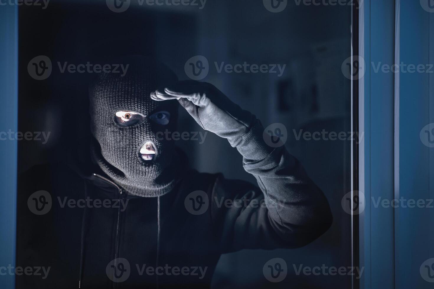 Portrait of housebreaker wearing black balaclava looking through window into residental building photo