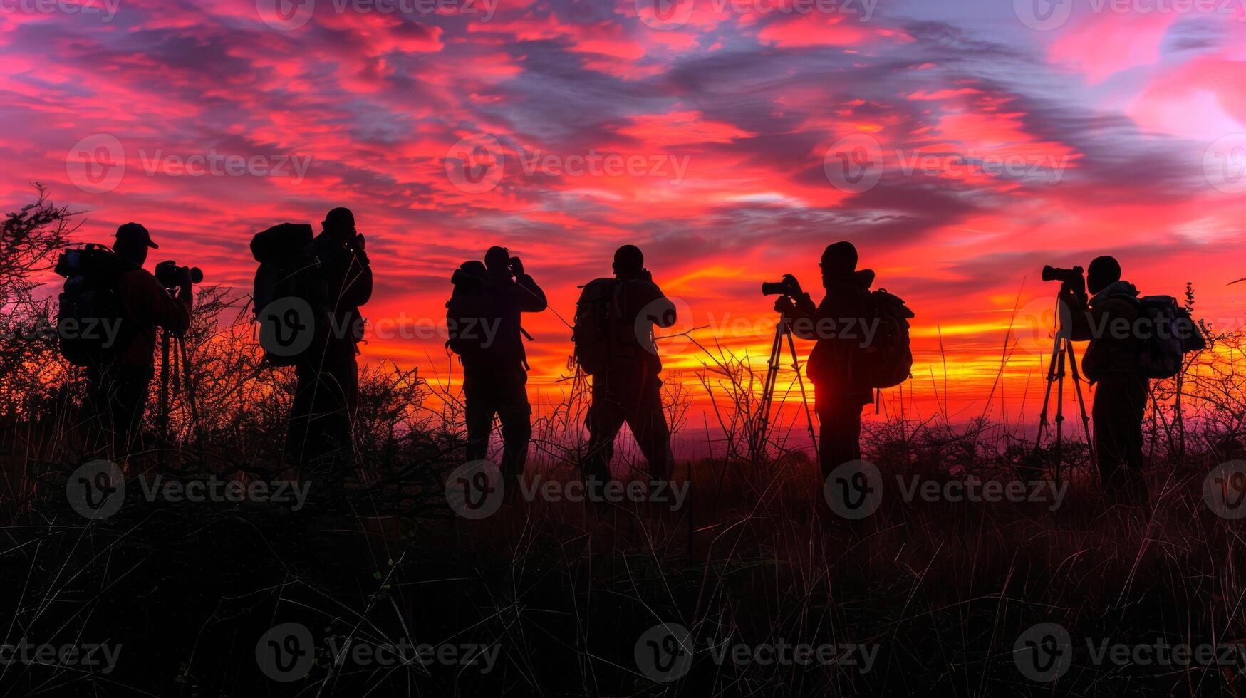 Groups of photographers stand on a hillside, silhouetted against a breathtaking sunset. The sky is ablaze with colors while they focus their cameras on the scenery. photo