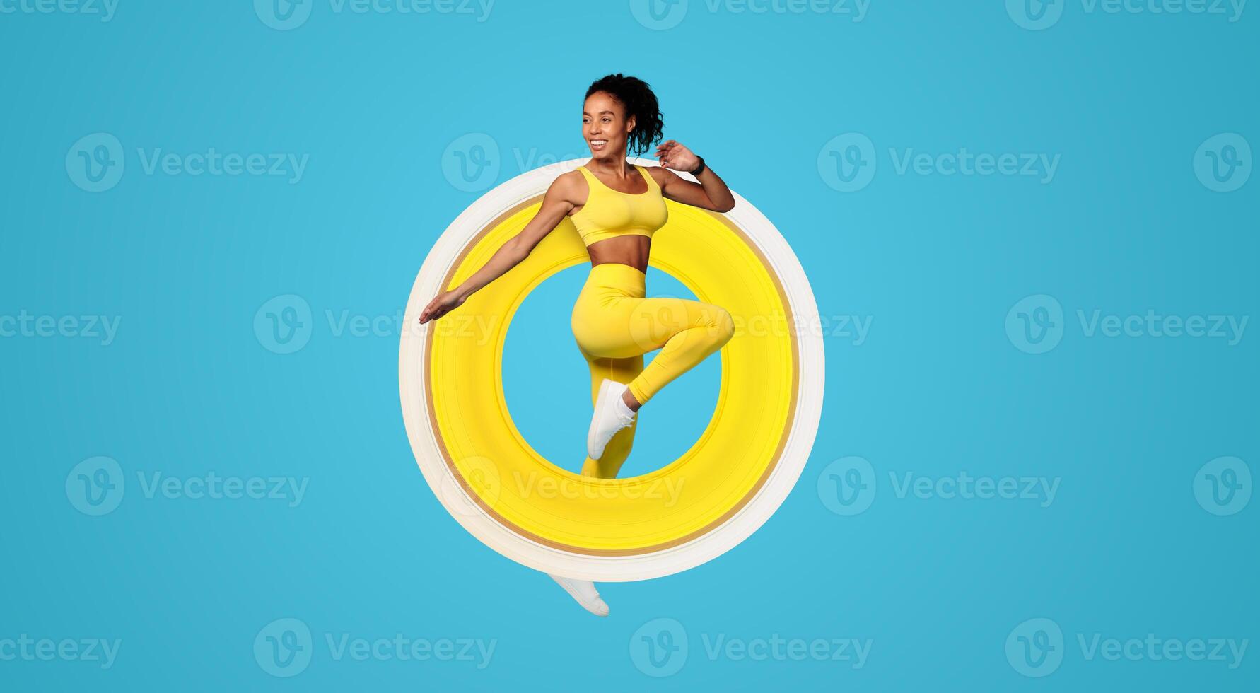 Young African American woman in yellow sportswear jumping doing knee to elbow exercise on blue studio backdrop, displaying athletic motivation during workout. Healthy sport training photo