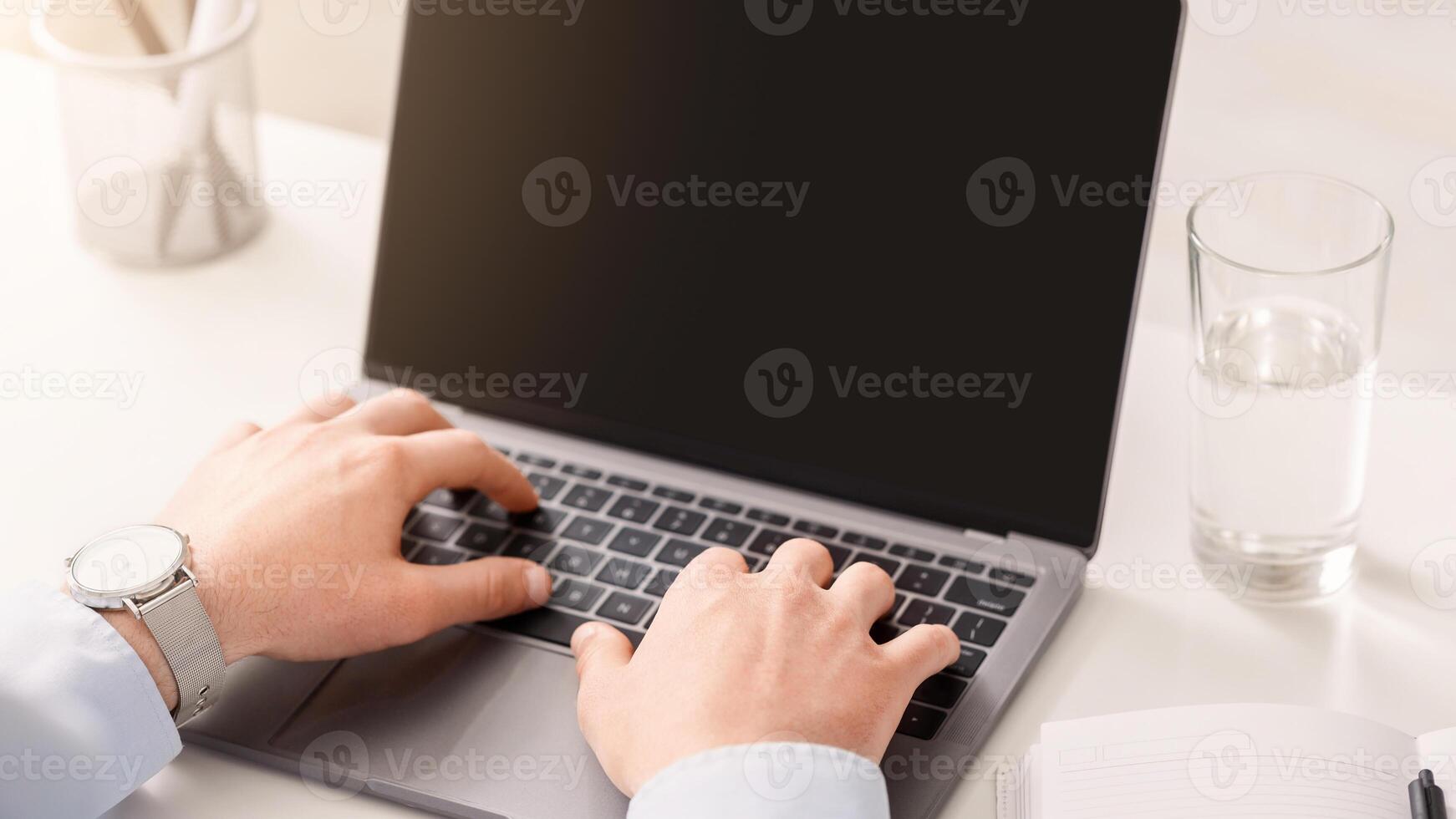 Man works at home. Hands of businessman typing on laptop with blank screen, on white table with glass of water photo