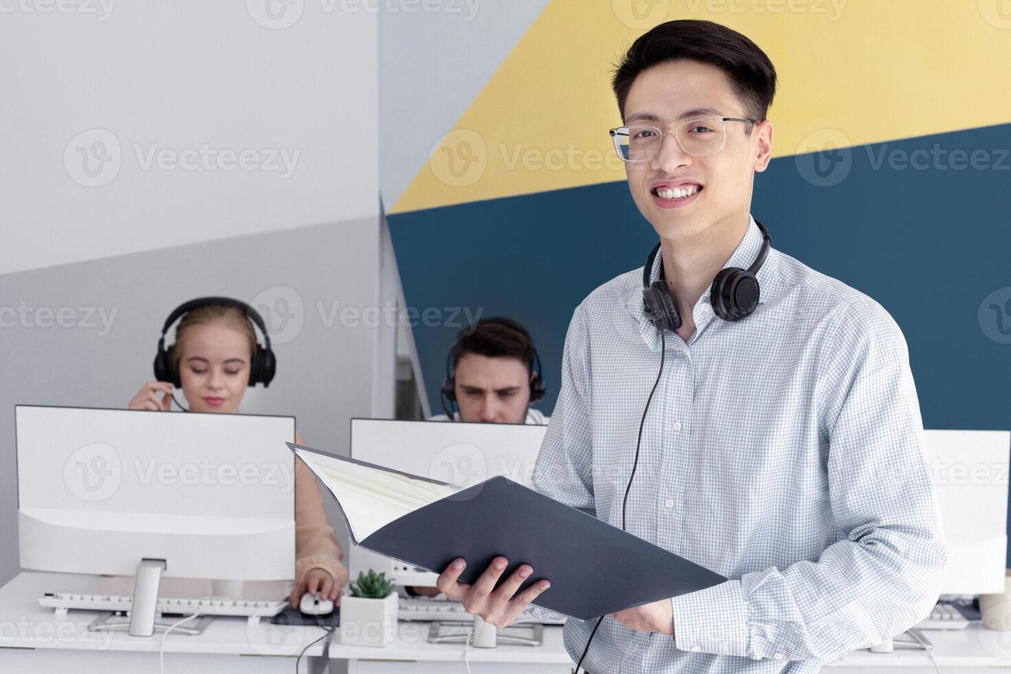 Happy Asian male helpdesk operator in headset holding documents at modern office. Portrait of smiling technical support consultant with coworkers at contemporary call centre photo