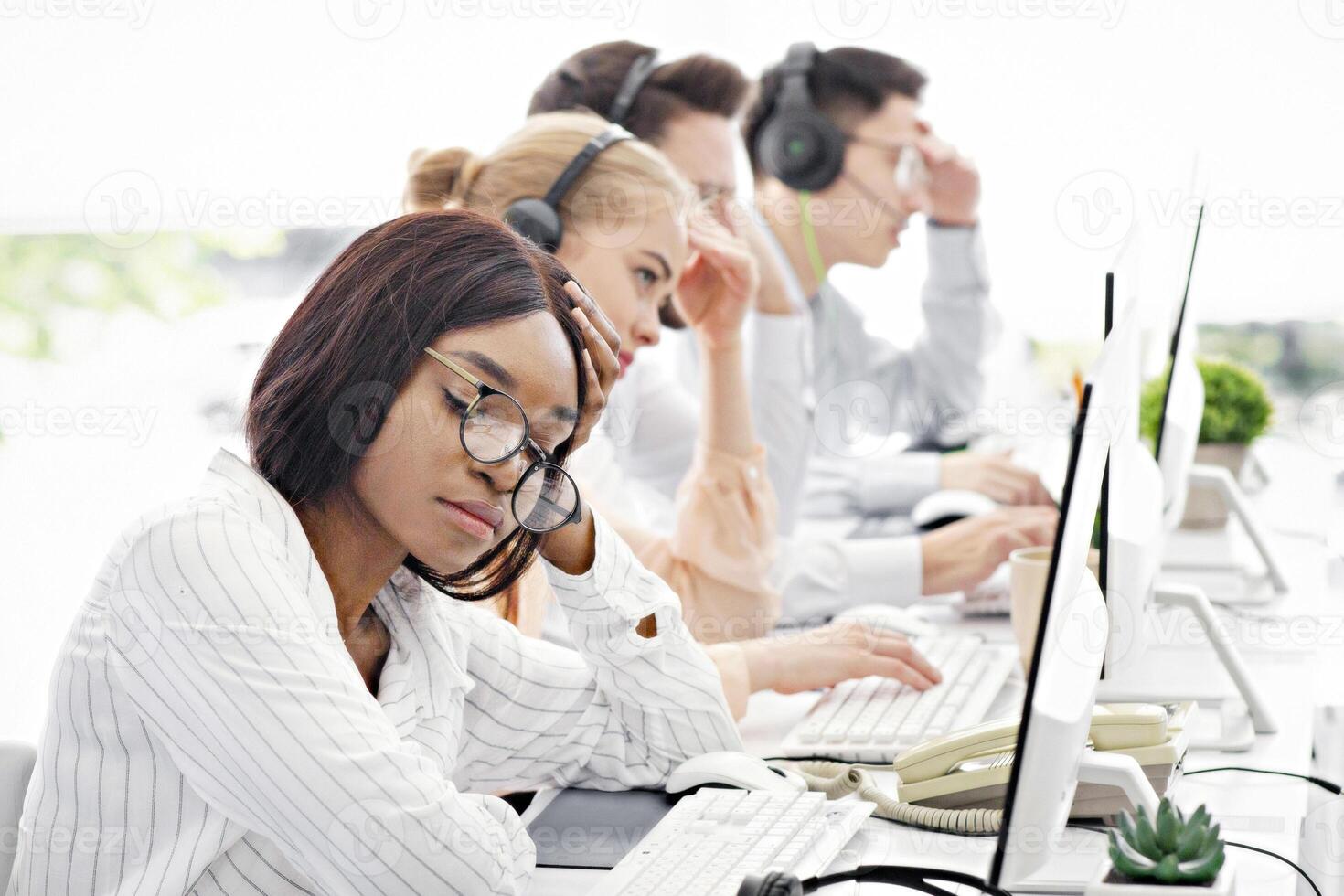 Overworked female helpline consultant procrastinating near computer monitor at call centre, blank space. Tired African American tech support agent feeling stressed at workplace photo