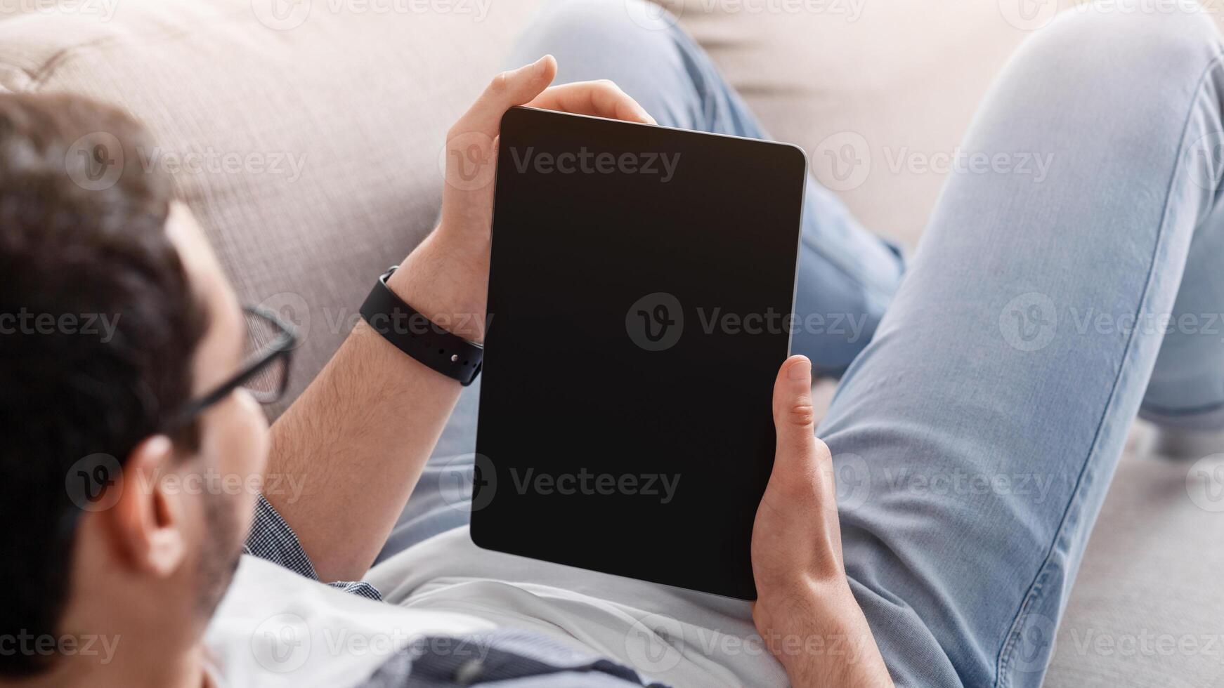 Reading books or blog and job search online. Young man with glasses lies on sofa and looks at tablet with blank screen photo