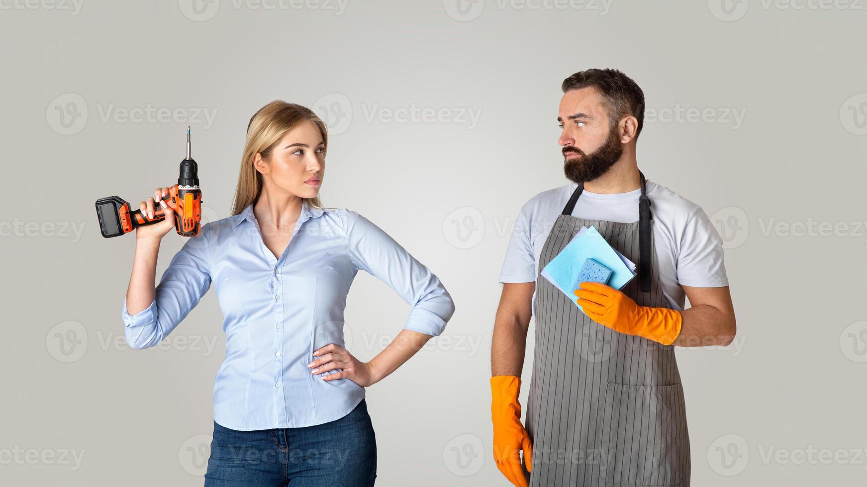 A woman stands confidently with a drill, facing a man holding a cloth, hinting at tension in their domestic relationship. Both are dressed for a home repair task at a neutral background. photo
